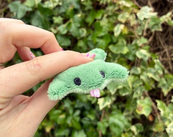 May include: A small, light green plush stingray toy with a soft, fuzzy texture. The stingray features two dark, embroidered circular eyes and a small, light pink ribbon tongue protruding from the center of its mouth area.