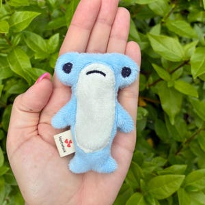 May include: A small, light blue plush hammerhead shark toy with a white belly, dark embroidered eyes, and a straight-line mouth. A small white fabric tag on the side features the text SophCraftsUK with three small red heart icons printed on it.