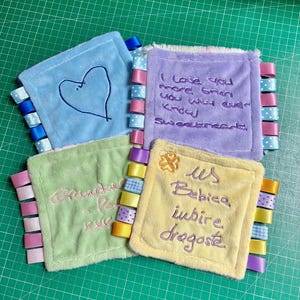 May include: Four square plush sensory blankets in light blue, lavender, light green, and pale yellow. Each features embroidered text or a heart shape and is bordered by colourful satin and patterned ribbon loops along the side edges.