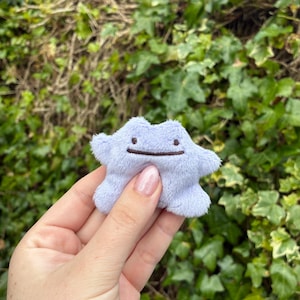May include: A small, light purple plush toy shaped like an amorphous blob with a simple embroidered face featuring two small dots for eyes and a straight horizontal line for a mouth. The soft, fuzzy material has a textured, velvety appearance.