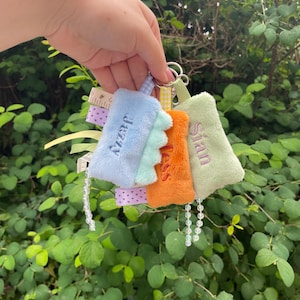 May include: A set of colourful plush keyrings, each with a name embroidered on them. The keyrings are in shades of blue, orange, and green, attached to a silver ring. Ribbons and beaded accents add detail. The background is a green bush.