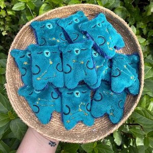 May include: A collection of small, teal plush bear-shaped figures arranged in a round woven basket. Each plush bear features embroidered closed eyes, a small nose, and a gold-stitched constellation pattern on its front surface.