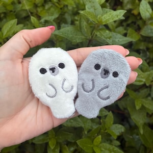 May include: Two small, plush seal-shaped patches, one white and one grey. Each patch features embroidered black eyes, a grey nose, and two curved grey lines representing flippers. The soft, fuzzy texture is visible on both oval-shaped figures.