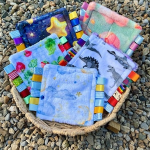 Könnte beinhalten: Eine Sammlung quadratischer sensorischer Decken aus weichem Plüschstoff mit verschiedenen Mustern wie Sternen, Erdbeeren, Dinosauriern und Wolken. Jede Decke ist mit mehreren bunten Bandschlaufen in verschiedenen Mustern und Farben versehen.