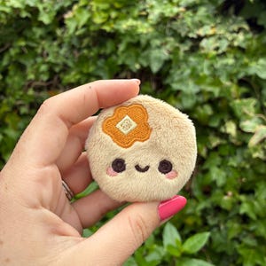 May include: A round, beige plush toy featuring an embroidered orange flower on the upper left side. The face consists of two dark brown circular eyes, a small curved mouth, and two light pink circular patches on the cheeks.