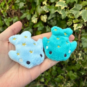 May include: Two plush whale-shaped toys in light blue and teal. Each has embroidered gold stars and black eyes. The toys are soft and fluffy, suitable for children or as decorative items.