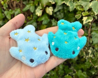 May include: Two plush whale-shaped toys in light blue and teal. Each has embroidered gold stars and black eyes. The toys are soft and fluffy, perfect for children or as decorative items.