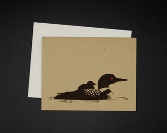 May include: A brown card with a black and white illustration of a loon with a chick riding on its back. The loon has red eyes.