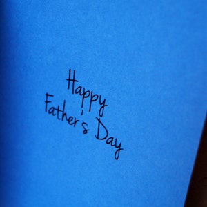 Puede incluir: Una tarjeta de felicitaci&oacute;n azul con el mensaje escrito a mano "Happy Father's Day" en escritura negra. La tarjeta est&aacute; abierta, revelando el mensaje en el interior. El fondo es de color neutro.