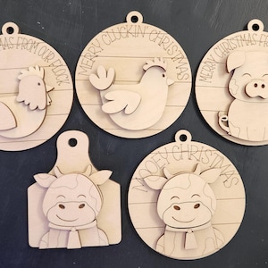 May include: A collection of five wooden Christmas ornaments featuring farm animals. Each ornament has a different animal: a rooster, a hen, a pig, a cow on a cutting board, and a cow in a circle. Each ornament has a festive message.