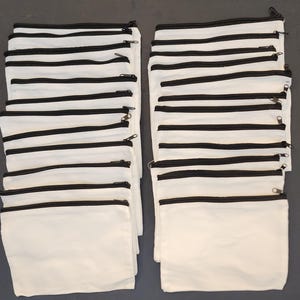 May include: A collection of white canvas zipper pouches with black trim and zippers. The pouches are rectangular and stacked in two groups, showcasing a simple, clean design. The black zippers provide a contrasting accent to the white fabric.