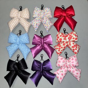 Pode incluir: Uma coleção de nove chaveiros em forma de laço em várias cores e padrões. Os laços incluem cores sólidas como vermelho, preto e roxo, bem como designs estampados com motivos florais e de confete. Cada laço tem um clipe de metal preto.
