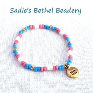 May include: A beaded bracelet with pink, white, blue, and gold beads. The bracelet has a gold charm with two horizontal lines. The text "Sadie's Bethel Beadery" is in the upper part of the image.