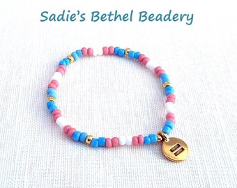Pode incluir: Uma pulseira de contas rosa, brancas, azuis e douradas. A pulseira tem um berloque dourado com duas linhas horizontais. O texto "Sadie's Bethel Beadery" é visível na parte superior da imagem.