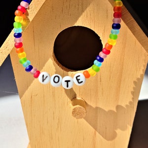 May include: A rainbow beaded bracelet with white letter beads spelling out the word "VOTE" on a wooden birdhouse.