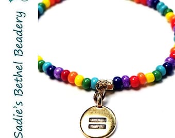 Pode incluir: Pulseira de contas de arco-íris com um berloque dourado com o símbolo de igual.