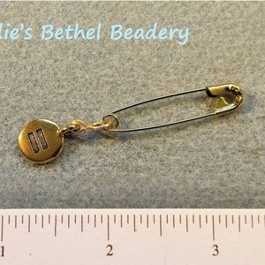 Pode incluir: Alfinete de seguran&ccedil;a dourado com um amuleto redondo com duas linhas paralelas. O amuleto &eacute; preso ao alfinete com uma pequena corrente. O alfinete tem aproximadamente 2,5 cm de comprimento.