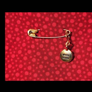 Pode incluir: Um alfinete de seguran&ccedil;a dourado com um pequeno amuleto dourado preso. O amuleto &eacute; redondo e tem duas linhas horizontais. O alfinete est&aacute; sobre um fundo vermelho com bolinhas brancas.