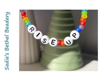 Pode incluir: Um bracelete de contas de arco-íris com contas de letras brancas que soletram "RISE UP".
