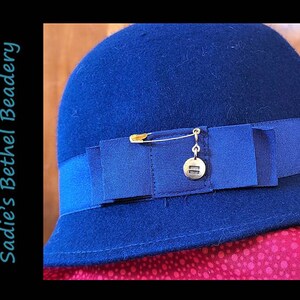 Pode incluir: Um chap&eacute;u de feltro azul marinho com aba larga e uma fita azul marinho em volta da coroa. Um alfinete de seguran&ccedil;a dourado com um pequeno amuleto dourado est&aacute; preso &agrave; fita.
