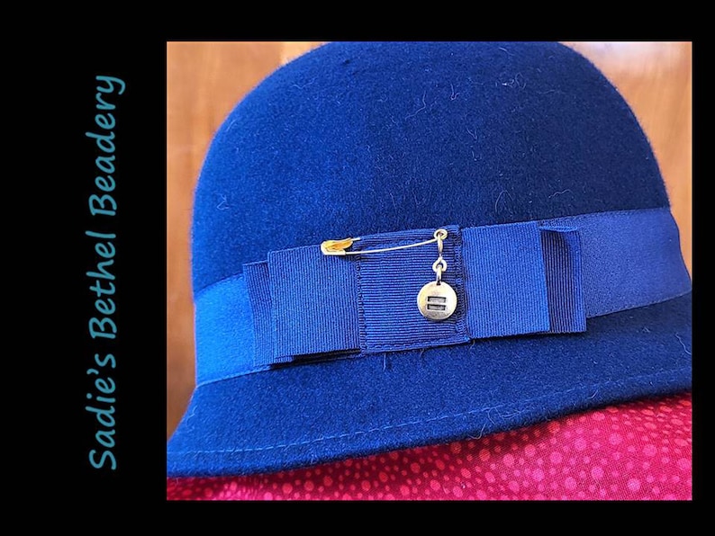 Pode incluir: Um chap&eacute;u de feltro azul marinho com aba larga e uma fita azul marinho em volta da coroa. Um alfinete de seguran&ccedil;a dourado com um pequeno amuleto dourado est&aacute; preso &agrave; fita.
