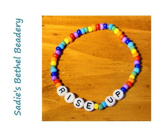 Pode incluir: Um bracelete de contas de arco-íris com contas de letras brancas que soletram "RISE UP".