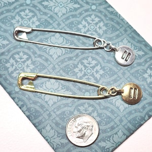 May include: Two safety pins with charms attached. The top pin is silver with a silver charm that has two parallel lines. The bottom pin is gold with a gold charm that has two parallel lines.