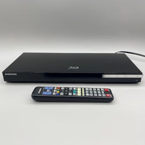 May include: Black Samsung Blu-ray player with a matching remote control. The player has a sleek, rectangular design with a silver trim and the Samsung logo. The remote has multiple buttons with various colors.