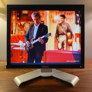 May include: A black Dell computer monitor displaying a scene from a television show featuring a person in a dark blazer playing an electric guitar, with a mural of a person holding a guitar in the background and the text BBC bbc.co.uk/doctorwho.
