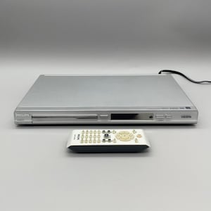 May include: Silver Philips DVD player with a matching remote control. The DVD player has a slot for discs and various buttons on the front panel. The remote is white with numerous buttons and a central navigation dial.