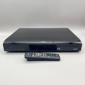 May include: A black Sharp Aquos Blu-ray player with a matching remote control. The player has the Aquos logo and the Blu-ray symbol on the front. The remote has multiple colored buttons.