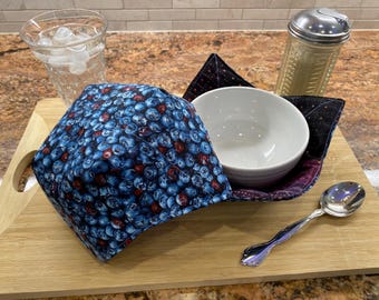 May include: A white ceramic bowl sits inside a blue and red blueberry-patterned bowl cozy. A silver spoon, a glass of water with ice, and a clear shaker are also on a wooden cutting board.