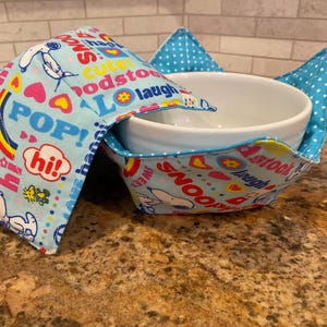 May include: A blue and white bowl cozy with a Snoopy and friends print. The cozy has a variety of words and phrases printed on it, including "Snoopy", "Pop", "Hi", "Sweet", "Happy", "Cute", "Goodstock", "Laugh", and "Best Look".