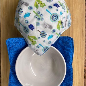 May include: A white ceramic bowl sits inside a blue patterned bowl cozy. Above, a matching bowl cozy with a white background features a pattern of blue, green, and gray birds and abstract shapes. The items are on a wooden cutting board.