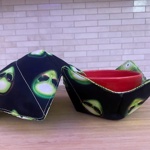 May include: Two fabric bowl cozies featuring a black background with a repeating pattern of green alien faces. One cozy is folded flat, while the other is open and holds a solid red ceramic bowl, showcasing the quilted construction and stitching.