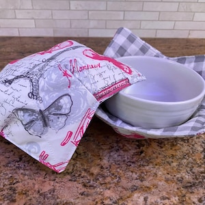 May include: A white ceramic bowl with a gray and white checkered fabric bowl cozy. The bowl cozy has a pink trim and a white fabric top with a black and gray butterfly and a Parisian-themed design with the text "Monsieur".