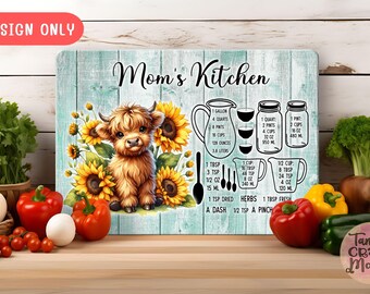 Könnte beinhalten: Dekoratives Küchenschild mit einer Highland-Kuh und Sonnenblumen. Der Hintergrund ist türkisblau mit dem Text "Mom's Kitchen" und Maßeinheiten. Frisches Gemüse ist um das Schild herum angeordnet.