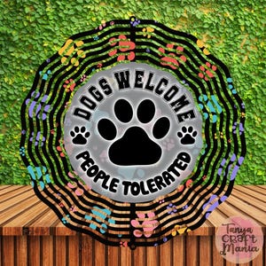 May include: A black metal sign with the text "DOGS WELCOME PEOPLE TOLERATED" and a large paw print. The sign has a circular design with colorful paw prints and wavy lines. The background is a green wall.