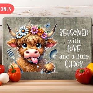May include: A brown and white Highland cow with a flower crown and a tongue sticking out. The cow is holding a spoon and a fork. The background is a gray textured surface with the text "Seasoned with love and a little chaos".