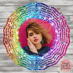 May include: A rainbow glitter wind spinner with a circular design. The wind spinner has a cutout in the center that is filled with a photo of a woman with brown hair. The wind spinner is on a wooden background.