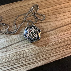 Op de afbeelding: Een zilverkleurige metalen ketting met een hanger die een cirkelvormig geometrisch levensbloempatroon toont over een donkere, veelzijdige basis. De hanger hangt aan een bijpassende zilverkleurige bolletjesketting met gedetailleerde uitsparingen.