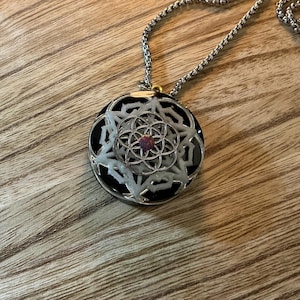 Op de afbeelding: Een ronde hanger aan een ketting met een zwarte basis en een wit, meerlaags geometrisch bloemenpatroon. Een kleine, iriserende paarse ronde steen is in het midden van het ontwerp geplaatst. De hanger hangt aan een zilverkleurige ketting.