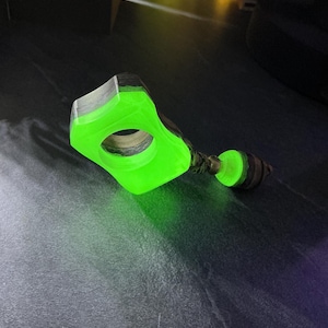May include: A bright neon green resin and dark wood finger ring featuring a geometric shape with a central circular opening. A small matching green resin and wood bead is attached to the side of the ring by a short, braided cord.