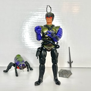 May include: A green and black action figure with a purple and black insect-like helmet and red eyes. The figure is holding a sword and has a black and green insect-like creature beside it.