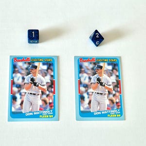 May include: Two baseball cards featuring Don Mattingly, with the text "Baseball's EXCITING STARS" and "FLEER '89". Two blue dice, one showing the number 1 and the other showing the number 2.