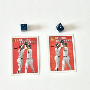 May include: Two baseball cards with the text "87 Record Breakers" and images of baseball players. Two blue dice with the numbers 1 and 2 are also present. The cards have a red border.