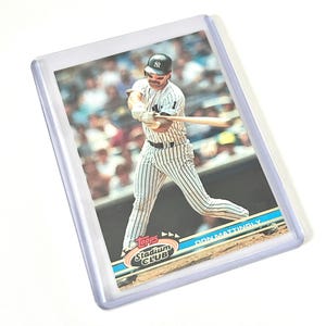 May include: A baseball card featuring Don Mattingly in a New York Yankees uniform, mid-swing. The card is from the Topps Stadium Club series and is encased in a protective sleeve.