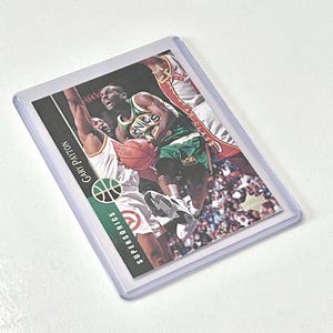 May include: A basketball trading card featuring Gary Payton of the Seattle SuperSonics. The card shows Payton in a green and white uniform, dribbling the ball while being guarded by a player in a red and white uniform. The card has a white background with black text and a basketball logo.