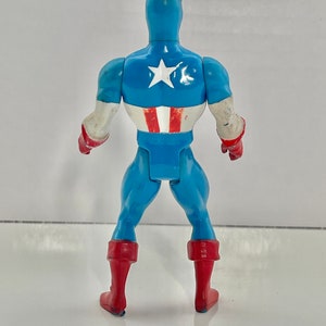 May include: A blue and red plastic action figure of Captain America. The figure is standing with its back to the camera. The figure has a red, white, and blue costume with a star on the chest.