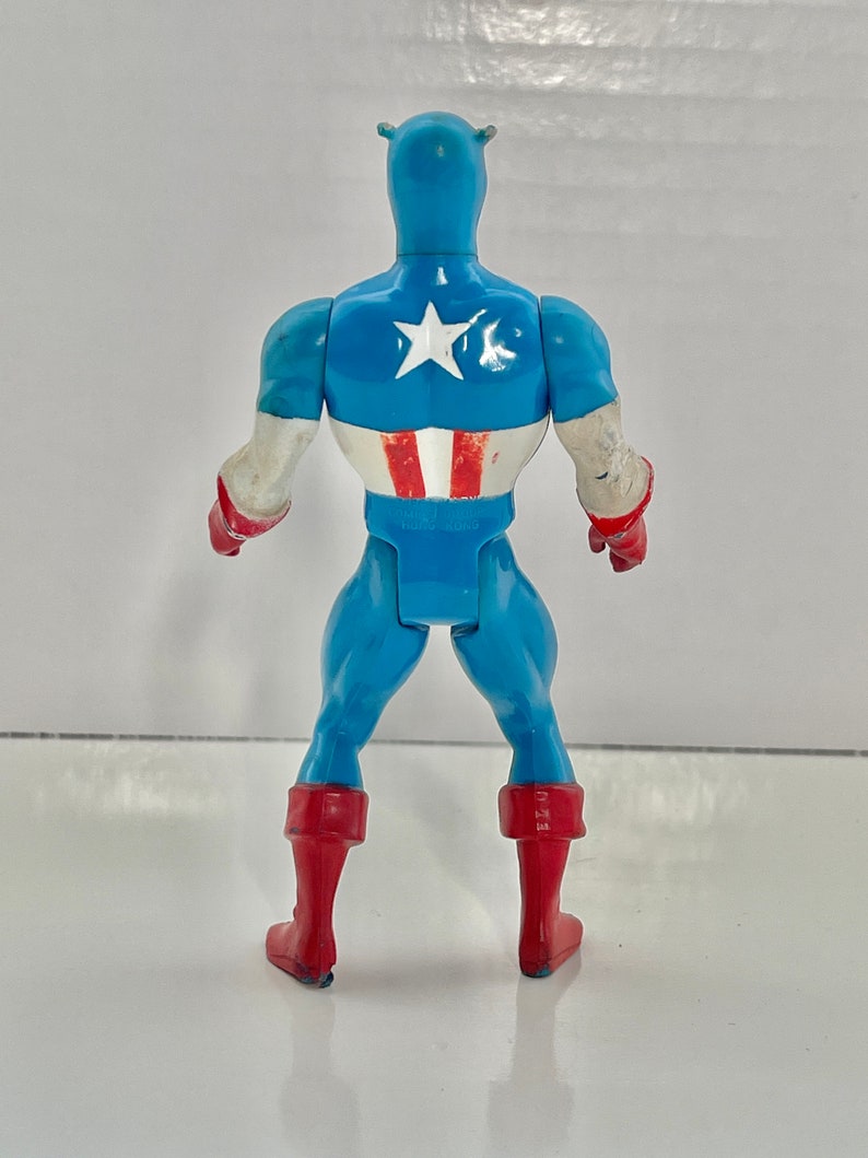 May include: A blue and red plastic action figure of Captain America. The figure is standing with its back to the camera. The figure has a red, white, and blue costume with a star on the chest.